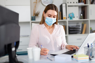 Fototapeta na wymiar Focused young woman in disposable face mask working in business office using laptop