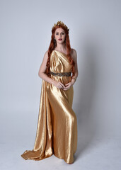 Full length portrait of girl with red hair wearing long grecian toga and golden wreath. Standing pose iisolated against a grey studio background.