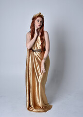 Fototapeta premium Full length portrait of girl with red hair wearing long grecian toga and golden wreath. Standing pose iisolated against a grey studio background.