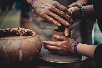 close up of hands of a potter and his student learning pottery at throwing wheel outdoors. Clay workshop. - obrazy, fototapety, plakaty