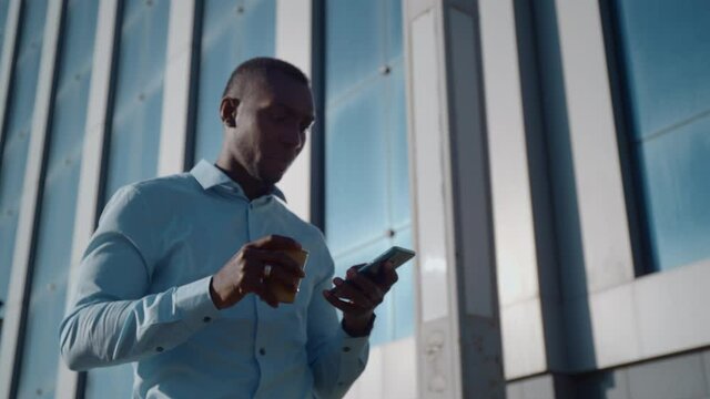 Successful African American business man walks down the street near a business office and uses a smartphone to check social networks.