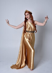 Full length portrait of girl with red hair wearing long grecian toga and golden wreath. Standing pose iisolated against a grey studio background.