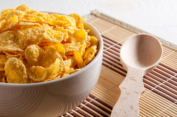 A white bowl of cornflakes, next to a wooden spoon. Everything is on a napkin on a white wooden table. Horizontal frame.
