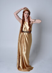Fototapeta premium Full length portrait of girl with red hair wearing long grecian toga and golden wreath. Standing pose iisolated against a grey studio background.