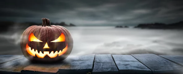 Keuken spatwand met foto One spooky halloween pumpkin, Jack O Lantern, with an evil face and eyes on a wooden bench, table with a misty gray coastal night background with space for product placement. © Duncan Andison