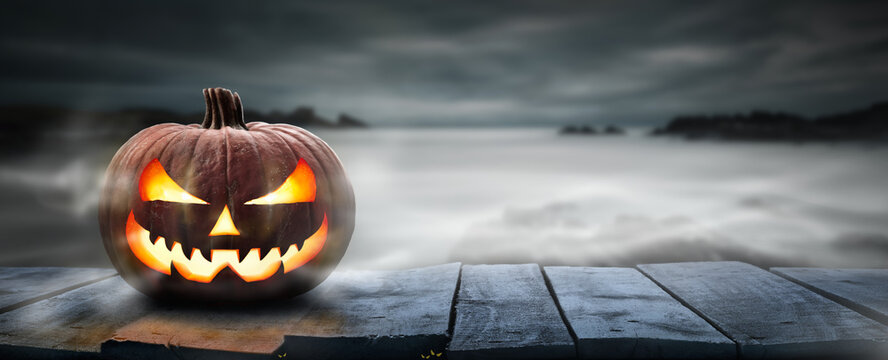 One spooky halloween pumpkin, Jack O Lantern, with an evil face and eyes on a wooden bench, table with a misty gray coastal night background with space for product placement.