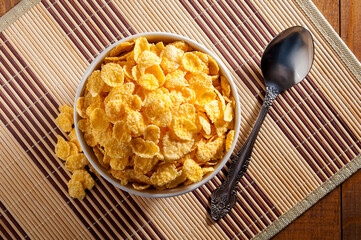 A white bowl of cornflakes, next to a metal spoon, all on a napkin that is on a brown wooden table. Composition in the center of the frame. Horizontal frame.