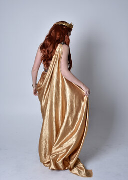 Full length portrait of girl with red hair wearing long grecian toga and golden wreath. Standing pose with back to the camera,  isolated against a grey studio background.