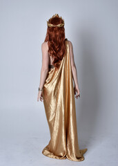 Full length portrait of girl with red hair wearing long grecian toga and golden wreath. Standing pose with back to the camera,  isolated against a grey studio background.