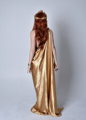 Fototapeta premium Full length portrait of girl with red hair wearing long grecian toga and golden wreath. Standing pose with back to the camera, isolated against a grey studio background.