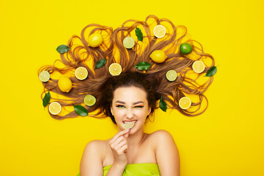 girl lying on yellow background with citrus fruits on long hair,young woman bites lemon wedge, food taste - Powered by Adobe