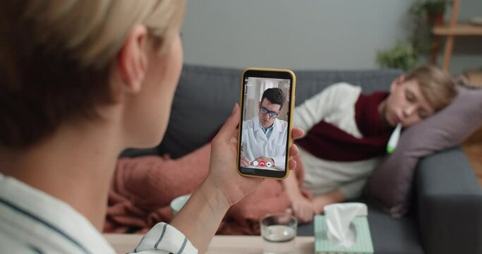 Overshoulder View Of Mother Holding Smartphone And Communicating With Male Doctor. Woman Having Online Medical Consultation While Her Ill Child Lying On Sofa. Concept Telemedicine