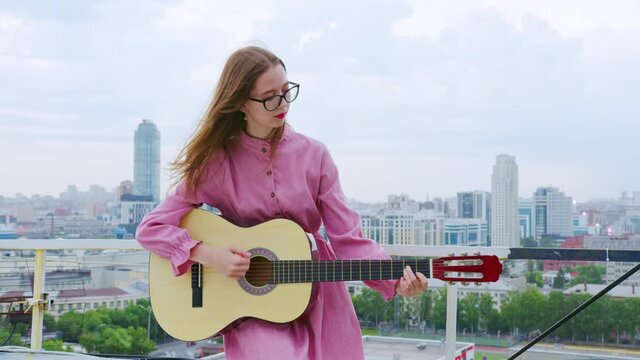 A young woman plays the guitar on the roof of a multistory building in the city. Evening time. The concept of creative people, freedom and loneliness