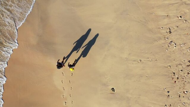 Top view tracking shot of two girls walking with their backpacks in slow motion as they go on an adventure at the beach during a bright sunny day nearby the waving sea.