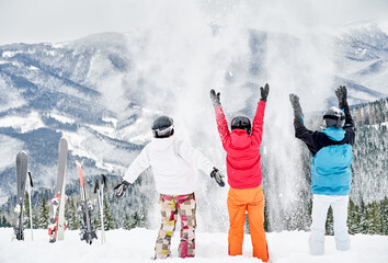 Back view of friends skiers in ski suits and helmets throwing fresh powder snow high in the air,...