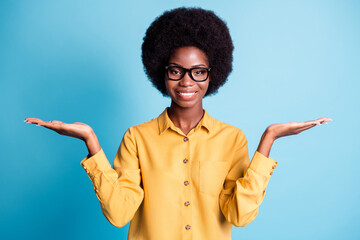 Photo of black skin big volume hairstyle girl beaming smiling hold two hands empty space suggest variants costumer free will wear specs yellow shirt isolated blue color background