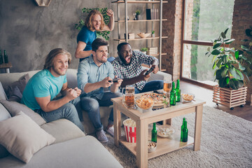 Full length photo of focused guy sit cozy couch play joystick want win two blonde hair pals support raise fists have table with crisps chips pint bottle in house indoors