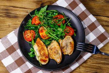 Fried chicken breast with salad of fresh arugula and cherry tomatoes on wooden table. Top view