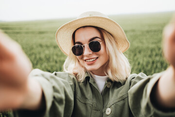 Portrait of Young Beautiful Girl Dressed in White T-shirt and Olive Jacket with Straw Holds Camera, Doing Selfie in the Green Field