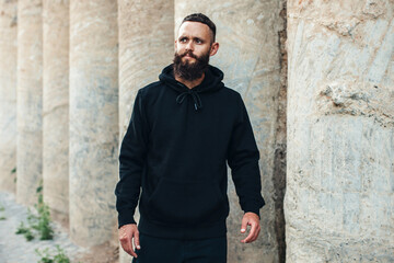 Obraz premium City portrait of handsome hipster guy with beard wearing black blank hoodie or sweatshirt with space for your logo or design. Mockup for print