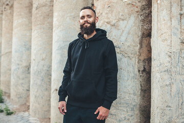 Fototapeta premium City portrait of handsome hipster guy with beard wearing black blank hoodie or sweatshirt with space for your logo or design. Mockup for print
