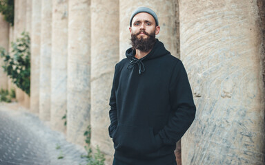 City portrait of handsome hipster guy with beard wearing black blank hoodie or sweatshirt with space for your logo or design. Mockup for print