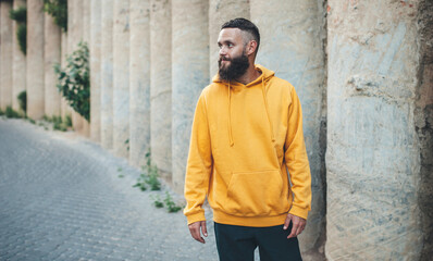 Fototapeta na wymiar City portrait of handsome hipster guy with beard wearing yellow blank hoodie or sweatshirt with space for your logo or design. Mockup for print