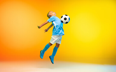Fototapeta na wymiar In jump. Young boy as a soccer or football player in sportwear practicing on gradient yellow studio background in neon light. Fit playing boy in action, movement, motion at game. Copyspace.