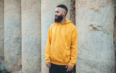 Obraz premium City portrait of handsome hipster guy with beard wearing yellow blank hoodie or sweatshirt with space for your logo or design. Mockup for print