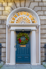 Fototapeta na wymiar Blue Georgian door, decorated for Christmas with green wreath and red decorations. Dublin, Ireland. Timber entrance with stone columns and decorative arch window fanlight
