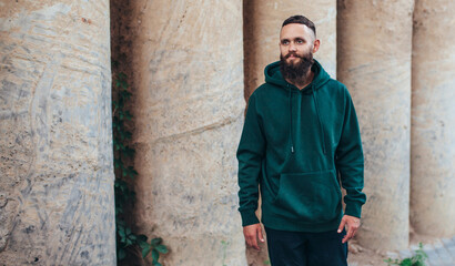 Fototapeta na wymiar City portrait of handsome hipster guy with beard wearing green (watercolor) blank hoodie or sweatshirt with space for your logo or design. Mockup for print