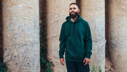 Fototapeta na wymiar City portrait of handsome hipster guy with beard wearing green (watercolor) blank hoodie or sweatshirt with space for your logo or design. Mockup for print