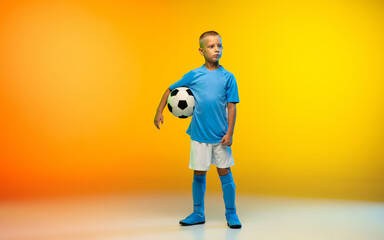 Fototapeta na wymiar Winner. Young boy as a soccer or football player in sportwear practicing on gradient yellow studio background in neon light. Fit playing boy in action, movement, motion at game. Copyspace.