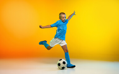 Attack. Young boy as a soccer or football player in sportwear practicing on gradient yellow studio...