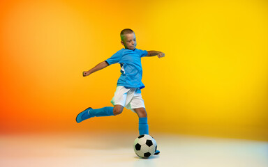 Kicking. Young boy as a soccer or football player in sportwear practicing on gradient yellow studio background in neon light. Fit playing boy in action, movement, motion at game. Copyspace.