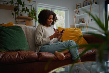 Happy young african woman using mobile phone while sitting a couch at home