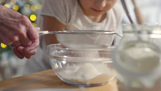 Handheld shot of unrecognizable elderly woman sieving flour into bowl as little girl watching. Fairy lights in background