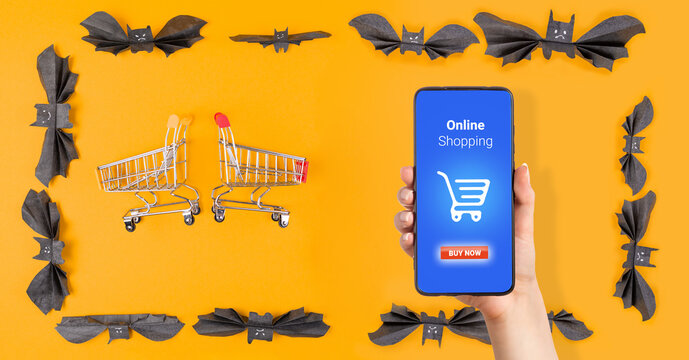 Two shopping carts and a woman's hand holding a smartphone are located in a frame of paper bats. Orange background. The concept of Halloween and online shopping