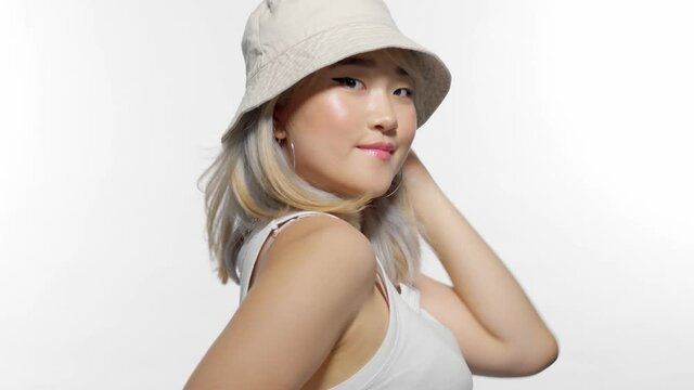 blonde Korean model in studio closeup portrait with bucket hat touches the hat and blowing hair makes v-sign hand and send kiss
