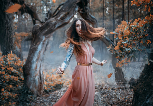 Fantasy portrait of a young red-haired woman in the autumn forest. The nymph girl runs and looks around. Long hair flying in the wind in motion. Background is nature dark trees, orange leaves falling