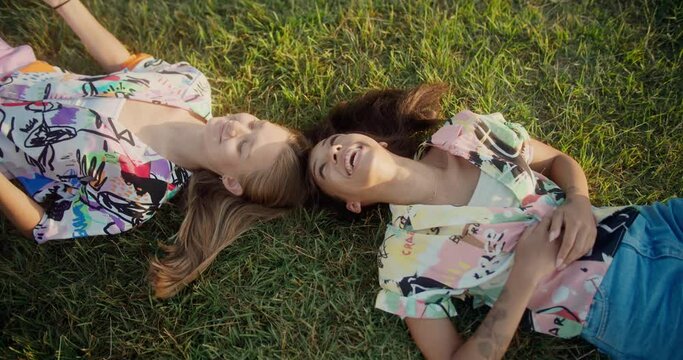 Shot from above of two positive pretty young female laughing having fun on green lawn, beautiful best friends enjoying summer sunny day smiling together in trendy outfits at grass in park outdoors