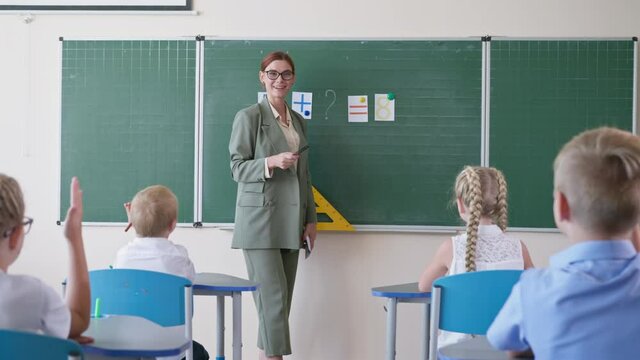 study mathematics, beautiful teacher in glasses at lesson near the whiteboard asks pupils questions, schoolchildren is ready to answer and raises hands