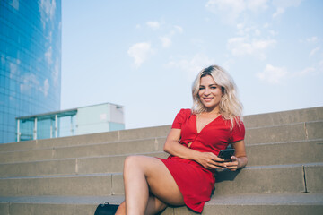 Portrait of happy Caucasian travel blogger resting at urban stairs enjoying free time for online networking via modern cellular device, millennial woman with digital smartphone smiling at camera