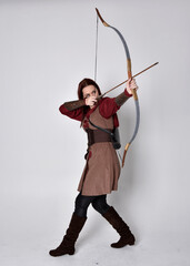 Full length portrait of girl with red hair wearing  brown medieval archer costume.. Standing pose holding a bow and arrow,  isolated against a grey studio background.