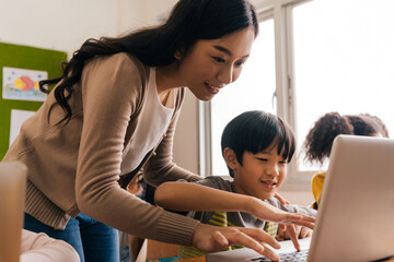 Young adult smiling beautiful Asian teacher helping elementary student boy with laptop in computer...