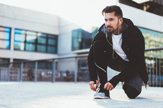 Determined Male Jogger In Tracking Suit Earphones Checking New Cross Sneakers For Jogging Prepare For Training Outdoors, Serious Caucasian Sportsman Listening Music And Tying Laces On Shoes