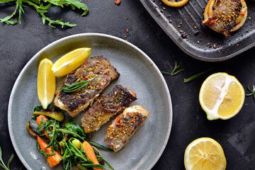 Grilled fish with lemon, herbs and rosemary. Fried fish fillets in a grey plate. Beautiful presentation of food. Rucolla salad and carrots. Dark background. Top view