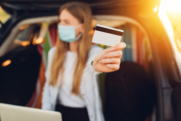 Happy young woman in a protective medical mask on her face, with a credit card uses a laptop, sitting in the trunk of a car against the background of the sunset, concept business, travel