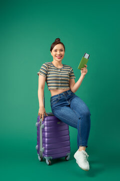 Attractive young Asian woman sitting on suitcase and holding passport, ticket flight over green background.