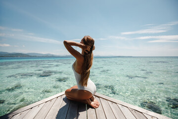 Woman in bikini at tropical beach sitting on her knees on wooden pier. Summer vacation concept....